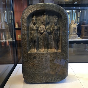 Ancient Egyptian stone stela carved with three standing deities and rows of hieroglyphs at the Louvre