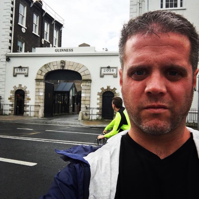 Selfie of a bearded man outside the arched Guinness Storehouse gate in Dublin as a cyclist passes