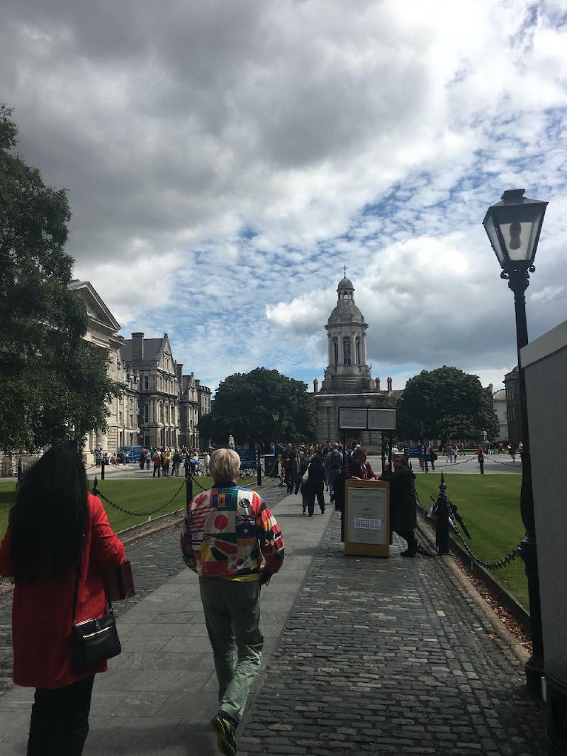 Pedestrians walk the cobbled path toward the Campanile at Trinity College Dublin, one in a jacket covered in world flags