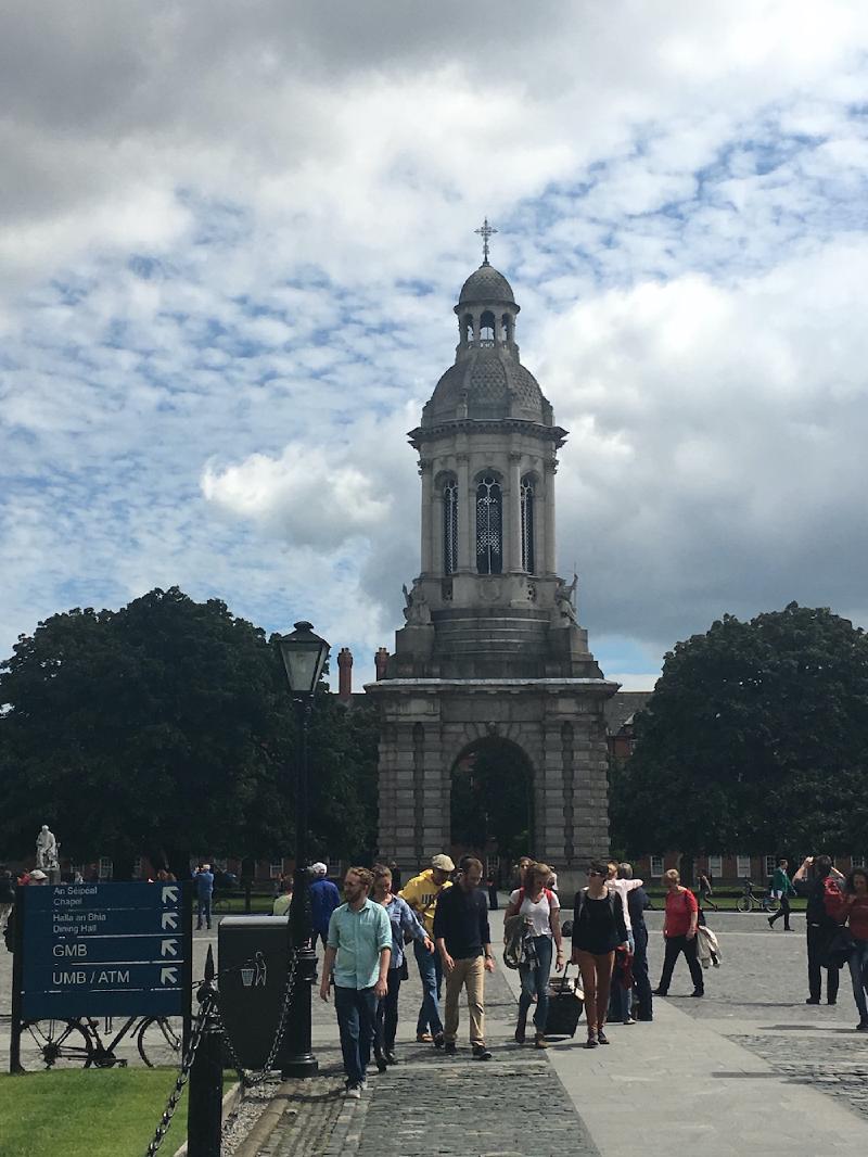 The stone Campanile bell tower at Trinity College Dublin, students strolling past campus signs below
