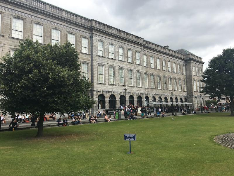 People rest along the long grey stone facade of Trinity College&rsquo;s Old Library beside a green lawn in Dublin