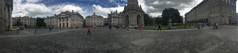 Panorama of a cobbled Dublin college square with a stone campanile, classical buildings, and people strolling under cloudy skies