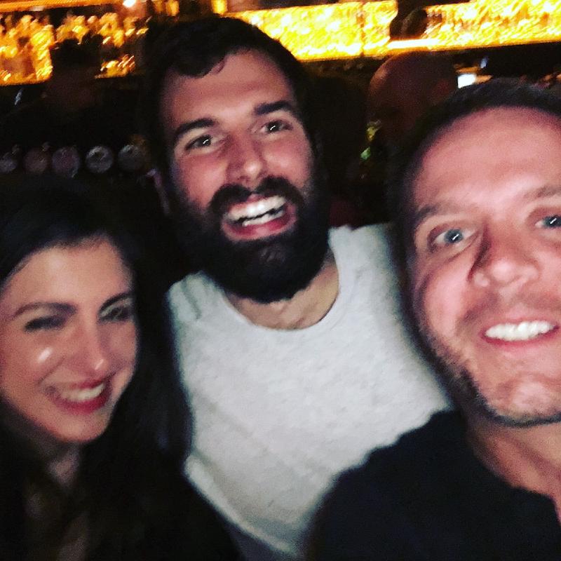 Three friends lean in smiling for a selfie in a dim Dublin bar with a glowing backlit bar behind them
