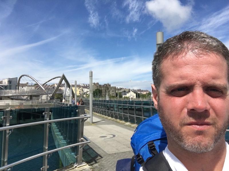 Man with a blue backpack takes a selfie beside a modern arched footbridge in a sunny harbor town
