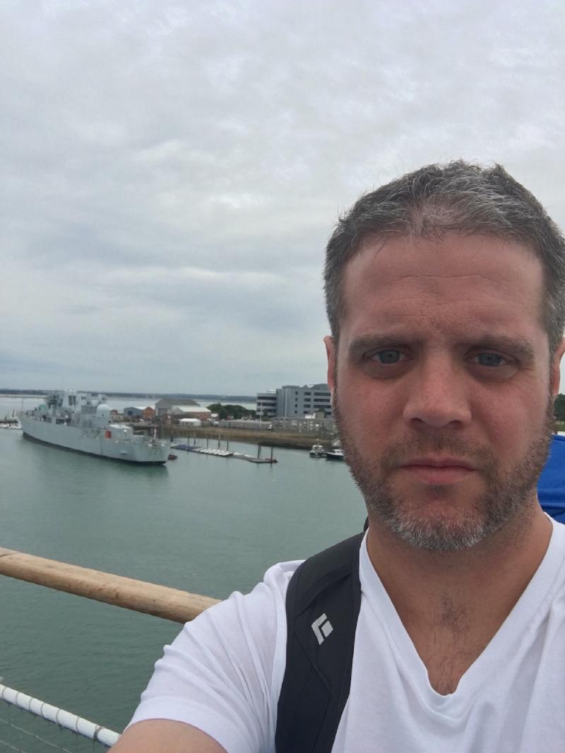 Selfie at a ferry railing with a gray navy warship moored across the harbor under an overcast sky
