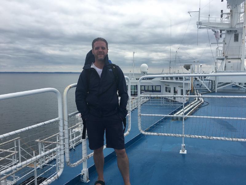 Man in a navy rain jacket and shorts stands on a ferry&rsquo;s blue upper deck under heavy gray clouds