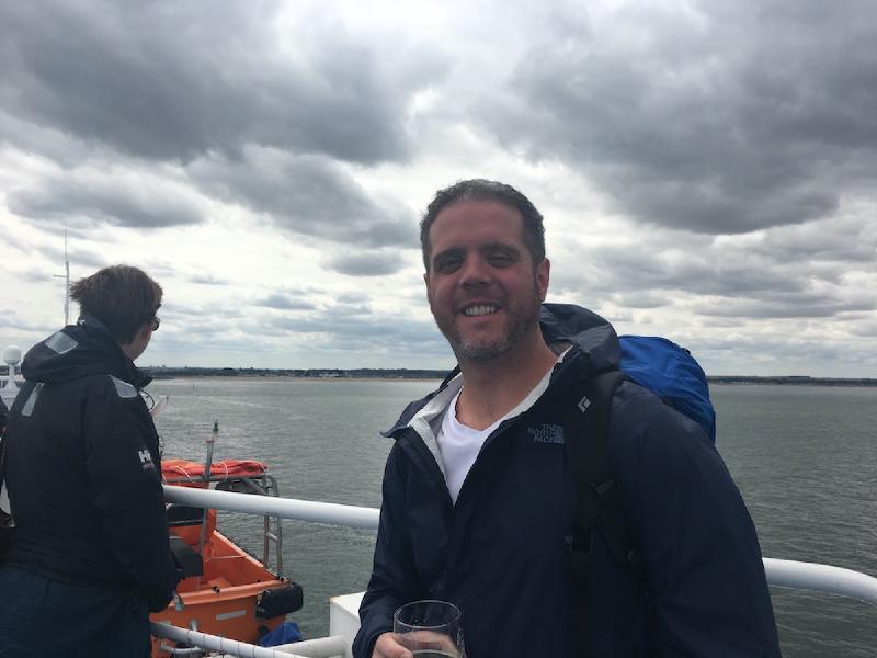 Smiling man in a navy jacket with a backpack holds a drink on a ferry deck, low coastline behind him