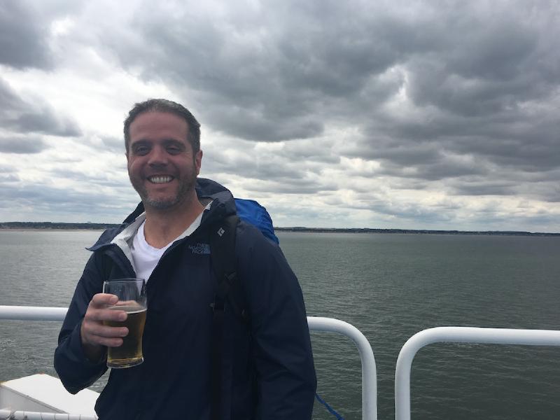 Man with a backpack grins while holding a pint of beer at a ferry railing under dark clouds at sea