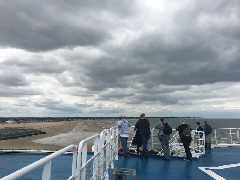 Passengers at a ferry deck railing watch a long sandy beach and low coastline pass under storm clouds