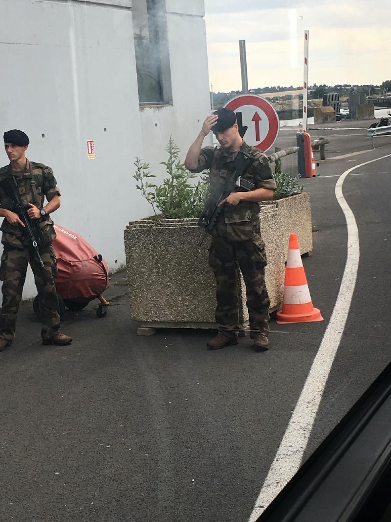 Two soldiers in camouflage with rifles stand guard beside a stone planter at a ferry-port checkpoint, seen from a vehicle