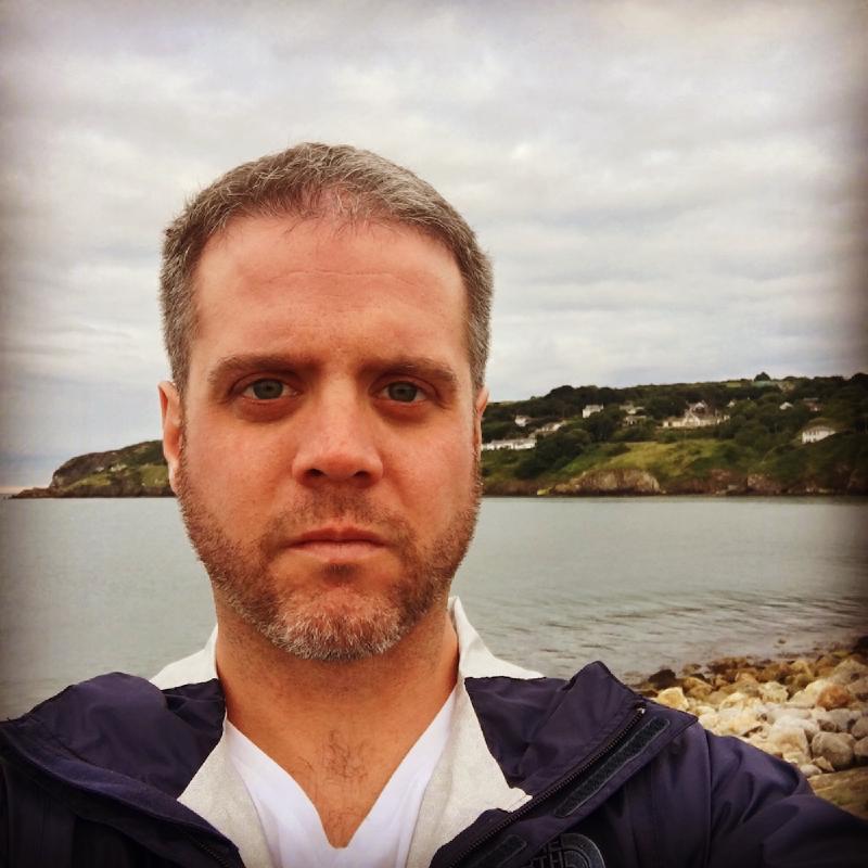 Close selfie on the rocky shore at Howth, with a headland of green cliffs and houses across the gray bay
