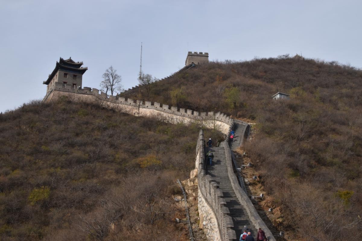 Stepped section of the Great Wall climbs a scrubby hillside toward a watchtower with a pagoda-roofed gate tower at left