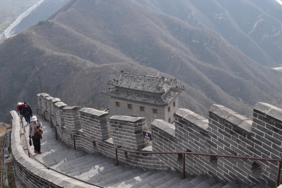 Curving battlements and steps of the Great Wall lead past a tiled-roof watchtower above hazy ridges