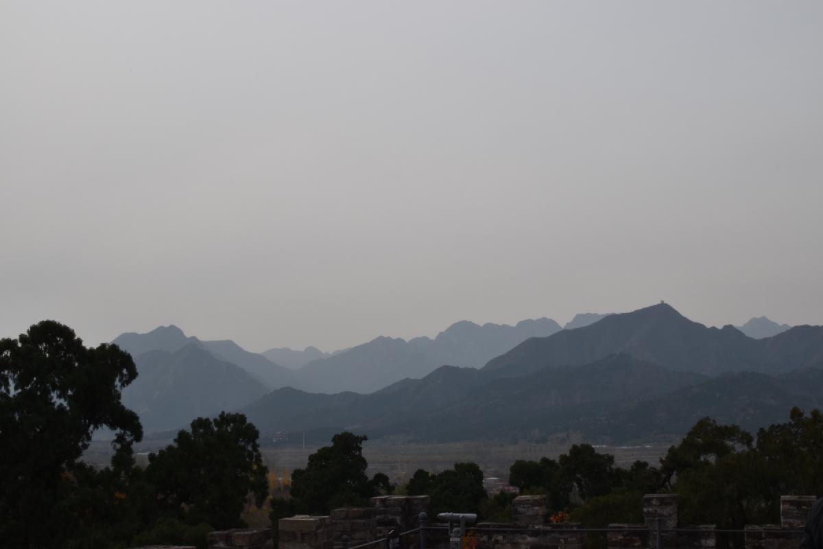 Hazy layers of mountain ridges fade into gray sky beyond dark treetops and Great Wall battlements