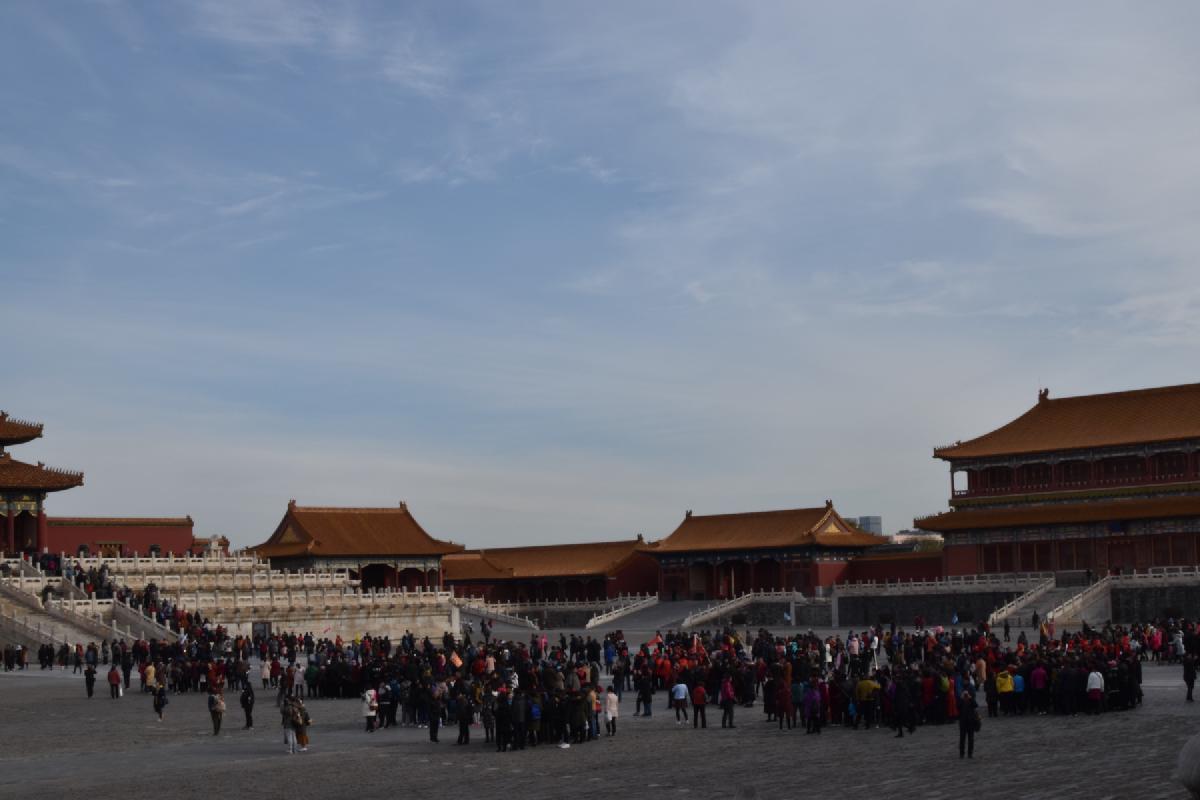 Tour groups gather in a vast stone courtyard of the Forbidden City ringed by red halls with golden roofs