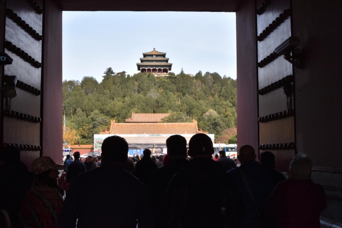 Silhouetted visitors exit a massive studded palace gate framing the hilltop pavilion of Jingshan Park