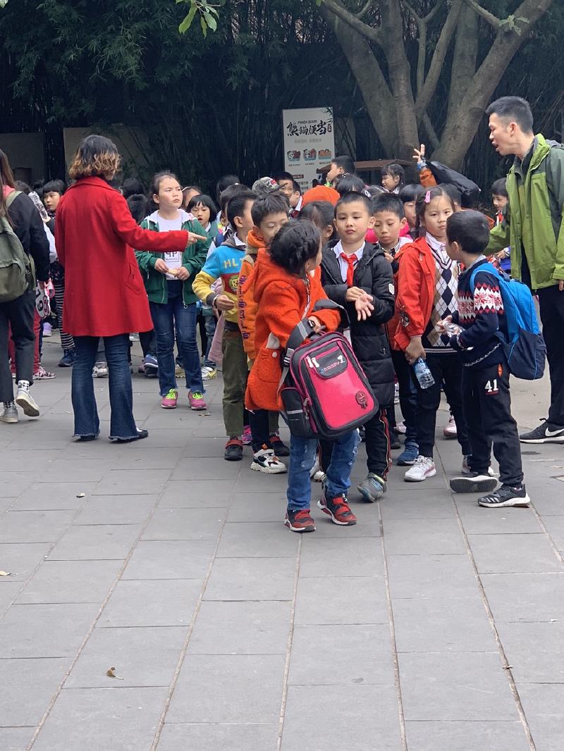 A school group of children with backpacks gathers on a paved plaza while adults direct them, Chengdu, China