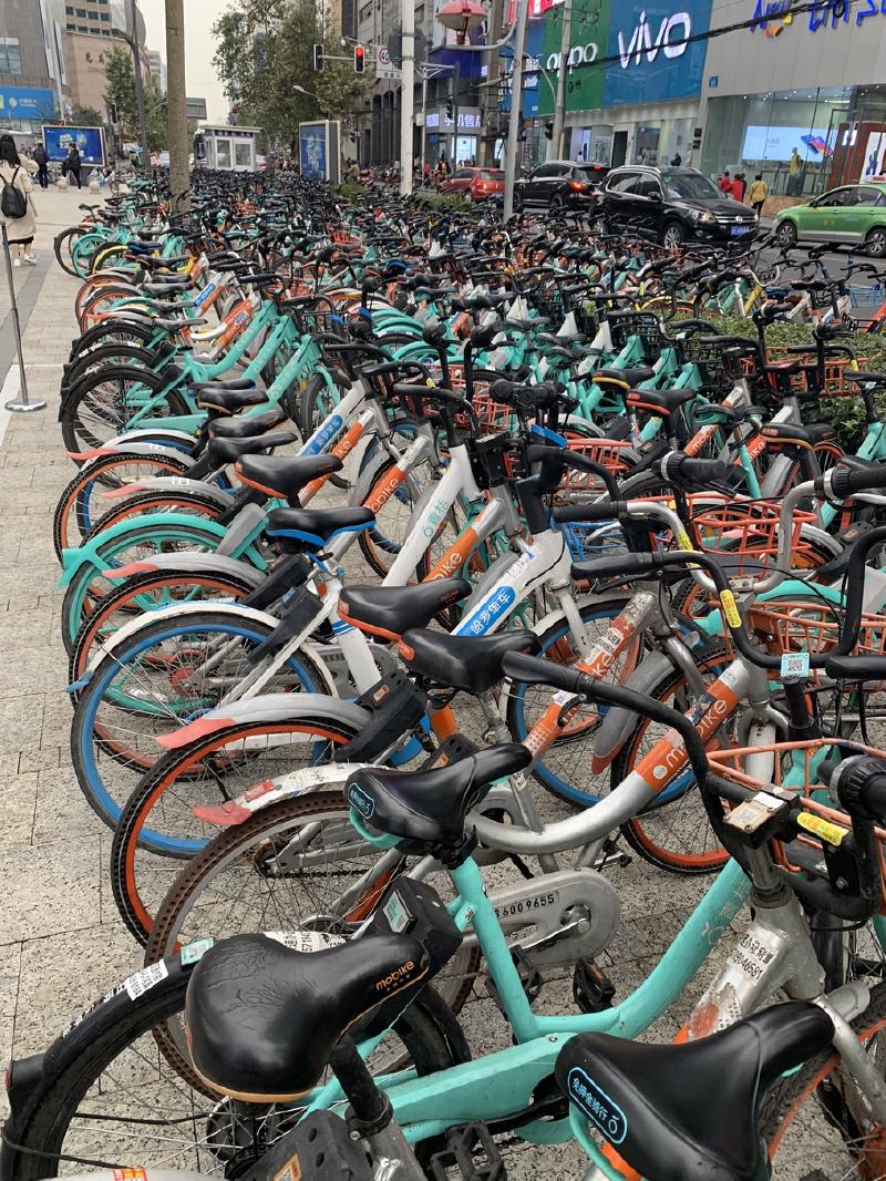 Hundreds of orange and teal share bikes packed along a city sidewalk below shop signs in Chengdu, China