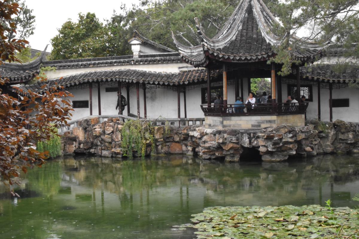 Visitors gather in a tiled pavilion above a rockery in a classical Chinese garden, its roof reflected in a lily-covered pond