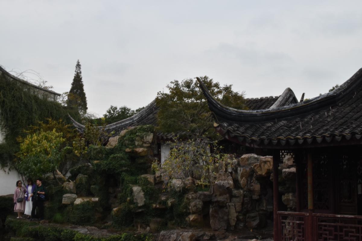 Three visitors stand at the foot of a garden rockery under curving tiled roofs and a grey sky in Shanghai