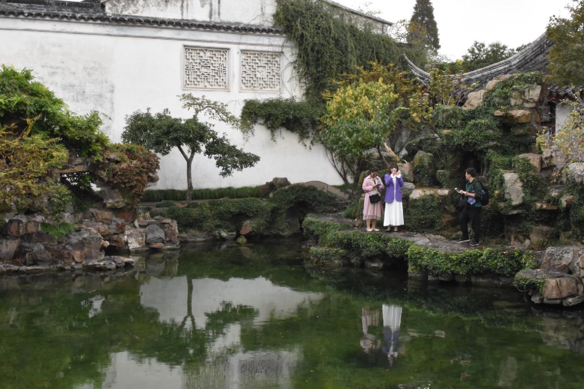 Two women taking phone photos stand on a mossy rock ledge above a still green pond, reflected below a white garden wall
