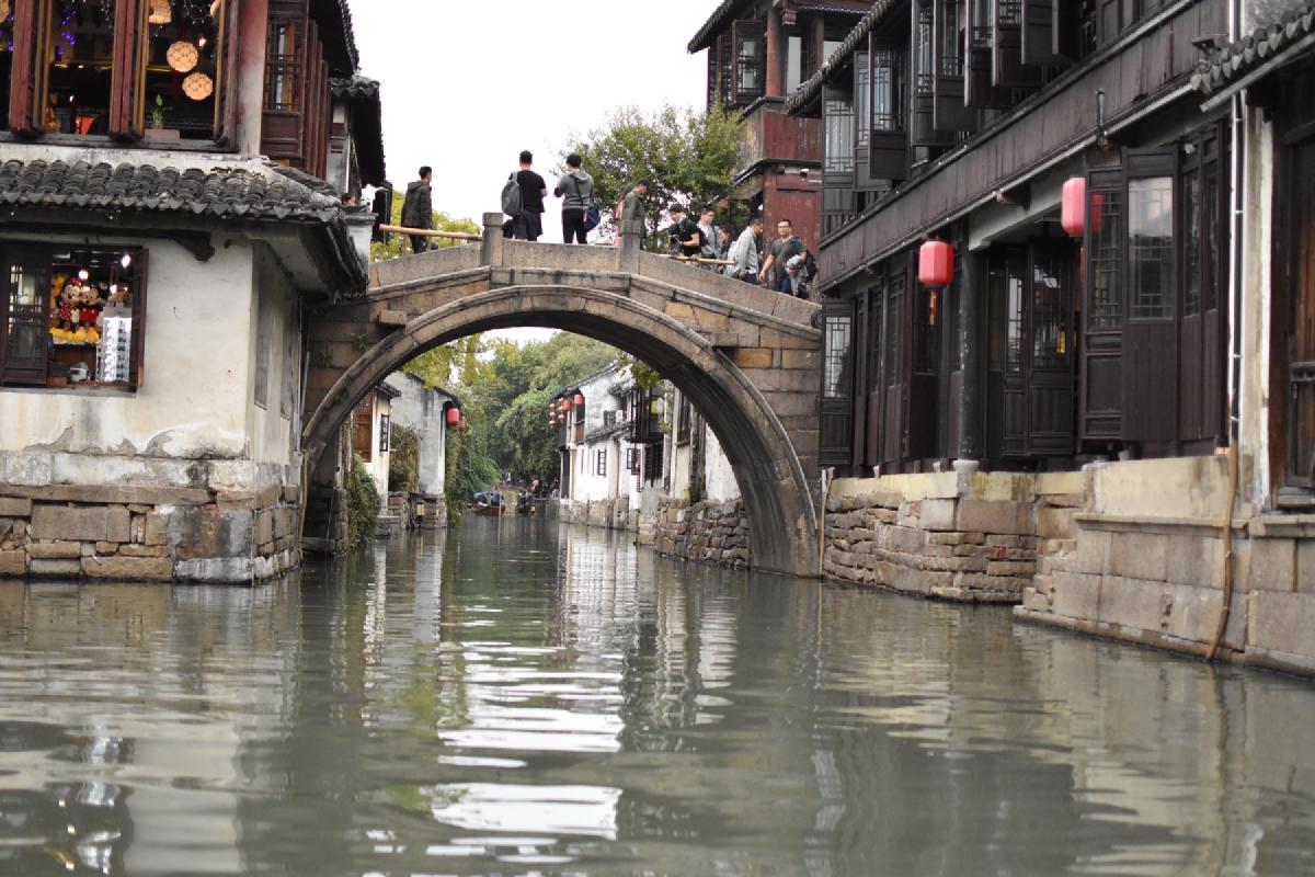 View from a boat of a stone arch bridge crowded with people, red lanterns hanging along the canal in China