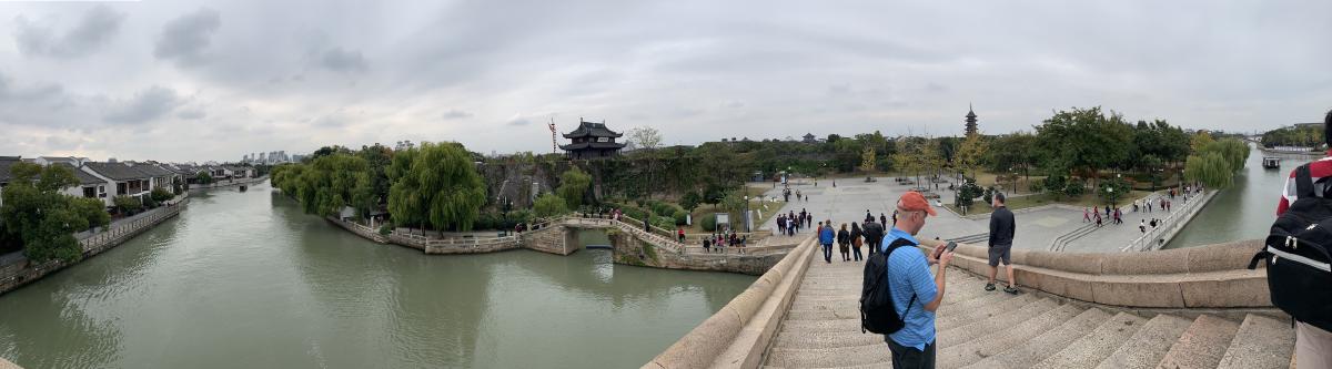 Panorama from a bridge over a wide canal in China, a man in an orange cap on the steps and a gate tower beyond