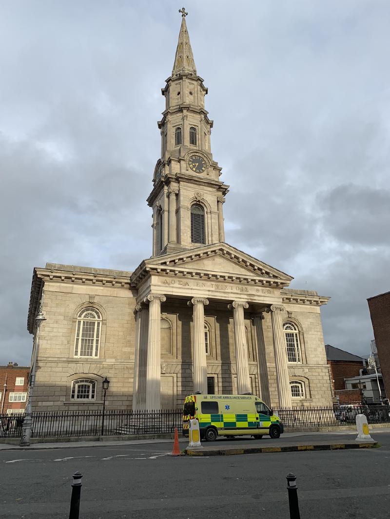 A columned stone church with a tall clock spire in Ireland, a yellow-green ambulance parked at its railings