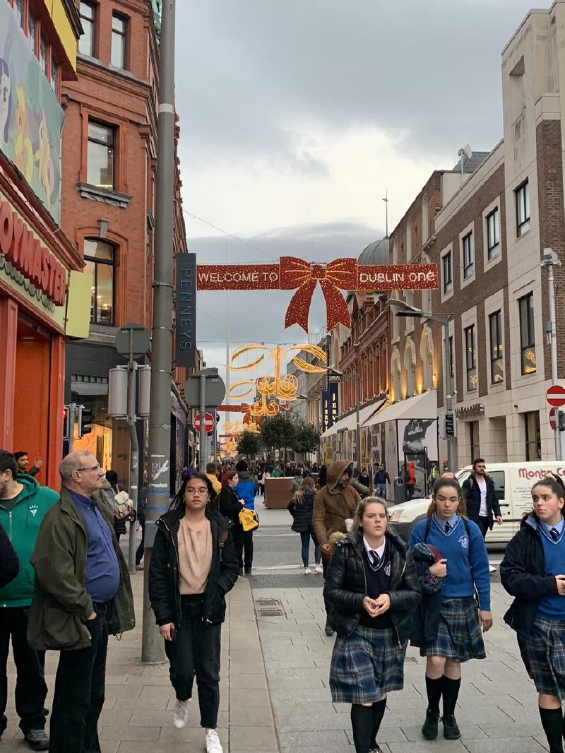 Shoppers and schoolgirls in uniform on a Dublin shopping street under a lit &ldquo;Welcome to Dublin One&rdquo; bow sign