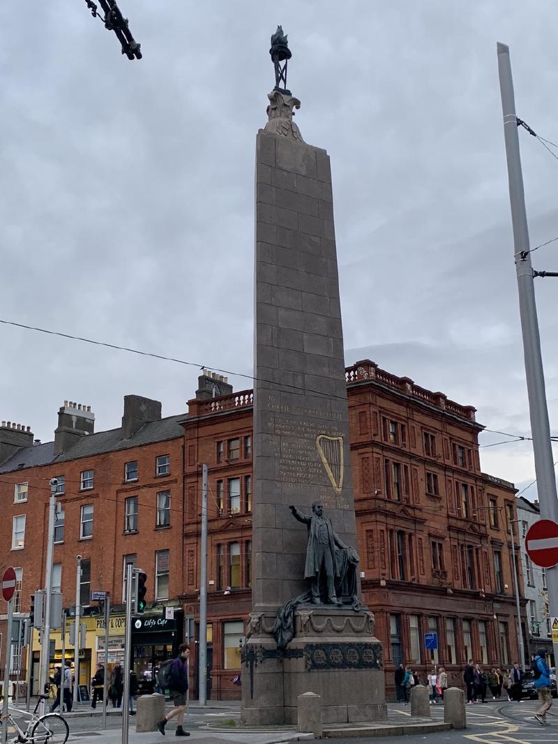 The Parnell Monument obelisk with its bronze statue and gold harp rises over a Dublin street corner under grey skies
