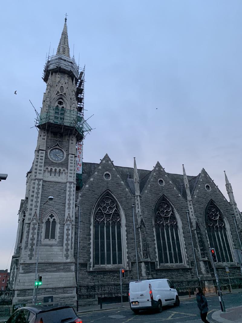 Stone Gothic church in Dublin with a scaffolded clock-tower spire, a white van passing on the street below at dusk