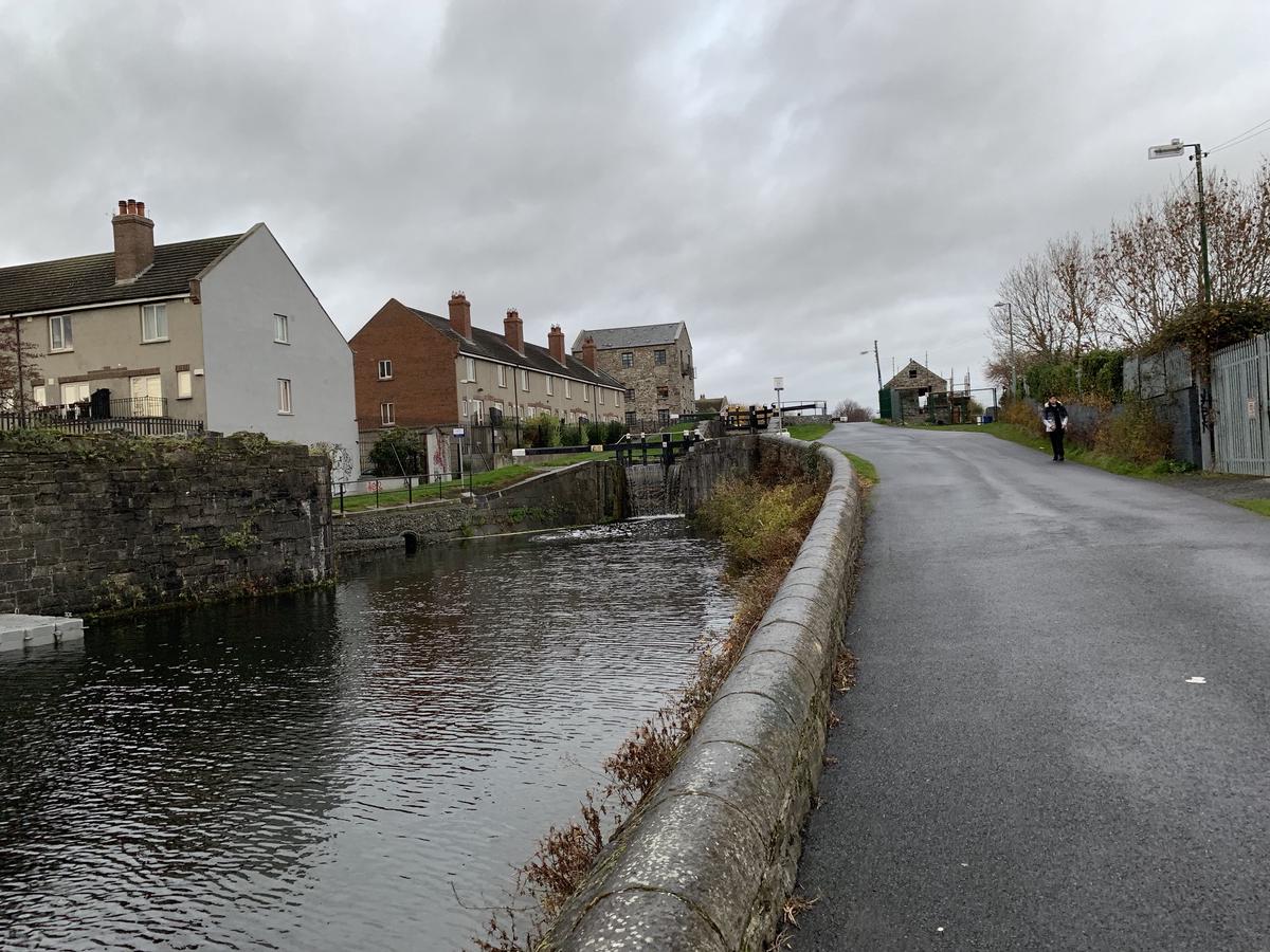 Canal lock with a small weir beside a lane in Dublin, brick houses on the far bank and a walker on the path