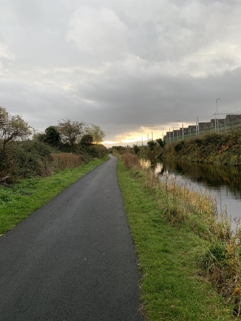 Paved canal towpath stretching toward a low sunset, hedges on the left and water with a green fence on the right