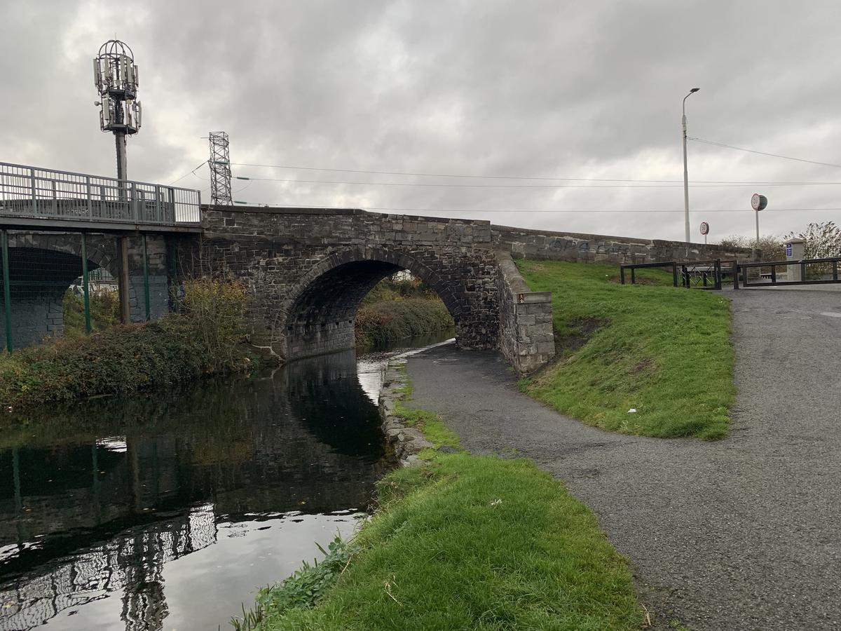 Stone arch bridge over a Dublin canal beside a modern rail bridge, with a towpath curving underneath on a grey day