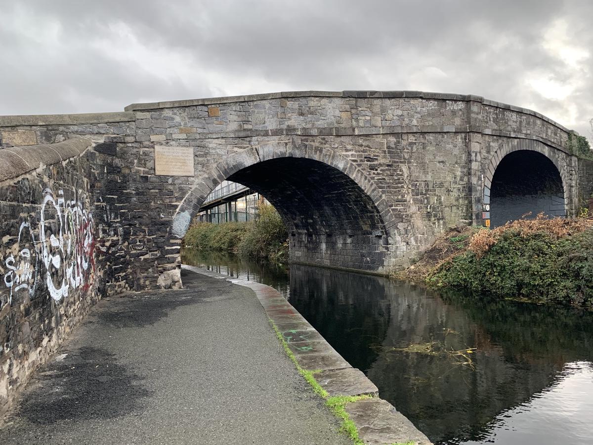 Graffiti-marked Broom Bridge arching over the Royal Canal in Dublin, a stone plaque set into the wall by the towpath