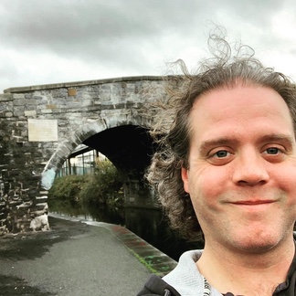 Curly-haired man taking a smiling selfie in front of Broom Bridge and the canal in Dublin