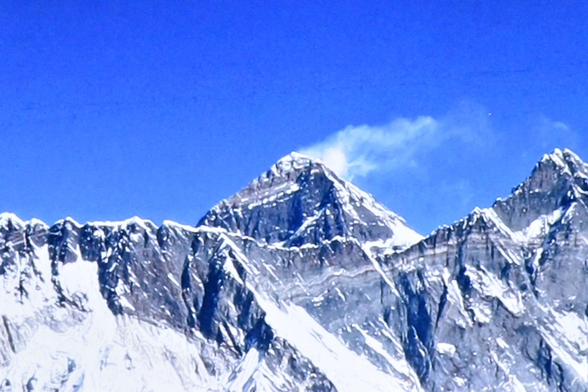 Snow plume streaming off the summit of Mount Everest against a deep blue sky, jagged ridges below