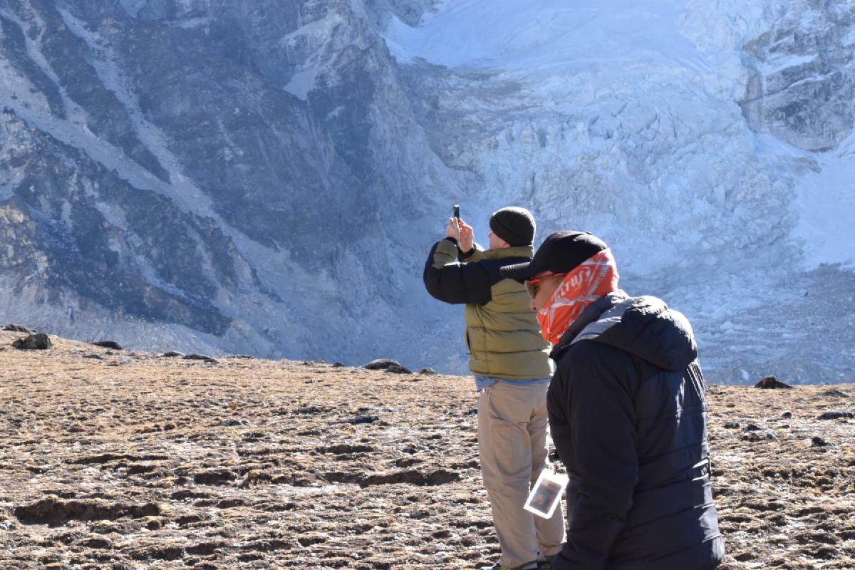 Two bundled-up trekkers on rocky ground in Nepal, one photographing a glacier spilling down the mountainside