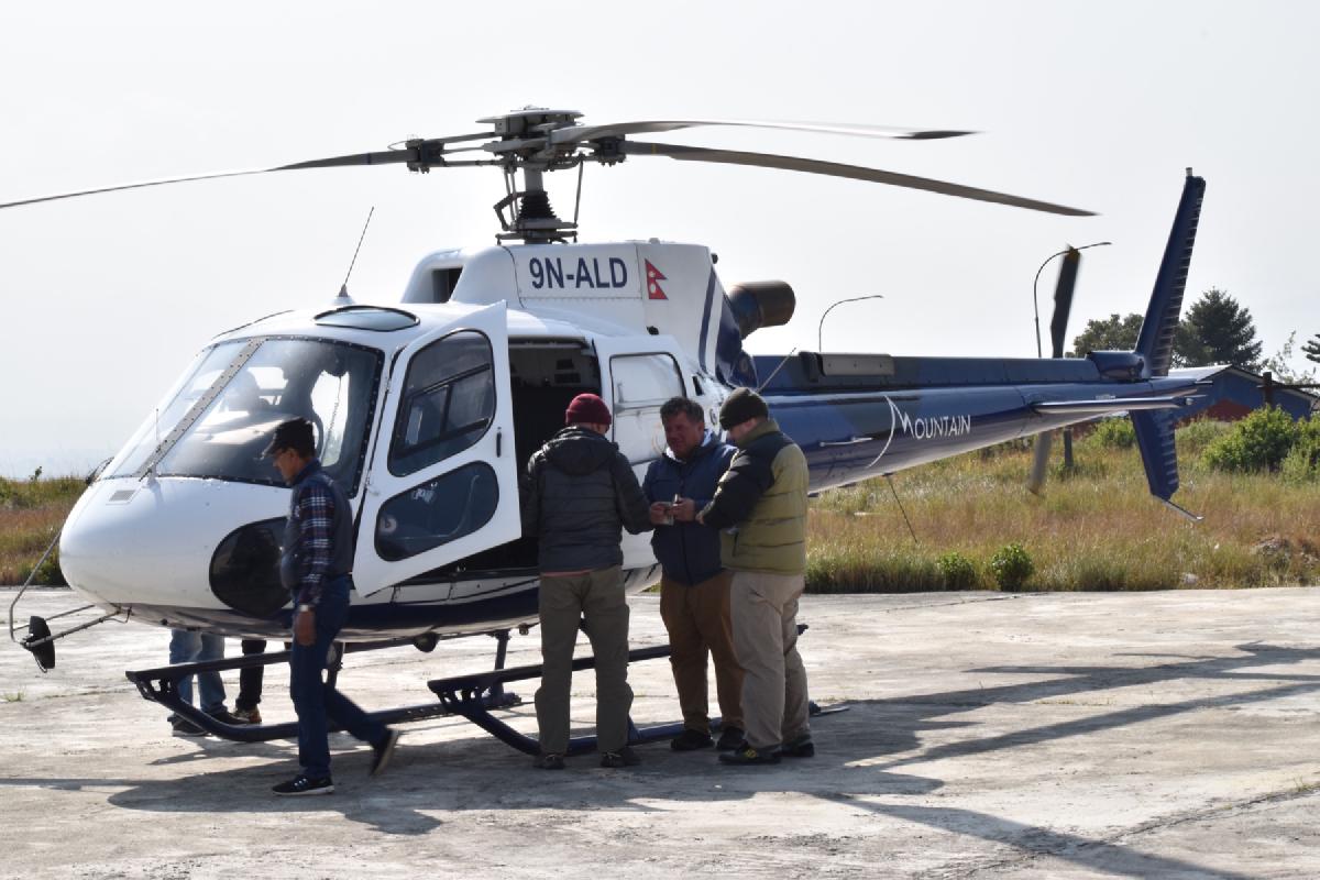 Group of trekkers gathered beside a white and blue helicopter on a concrete pad in Nepal, doors open before boarding