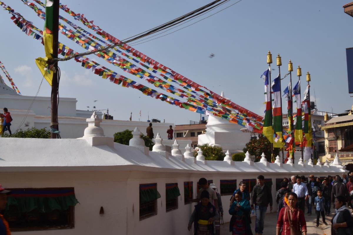 Strings of prayer flags fan out over a white wall lined with prayer wheels at Boudhanath in Kathmandu, visitors walking by