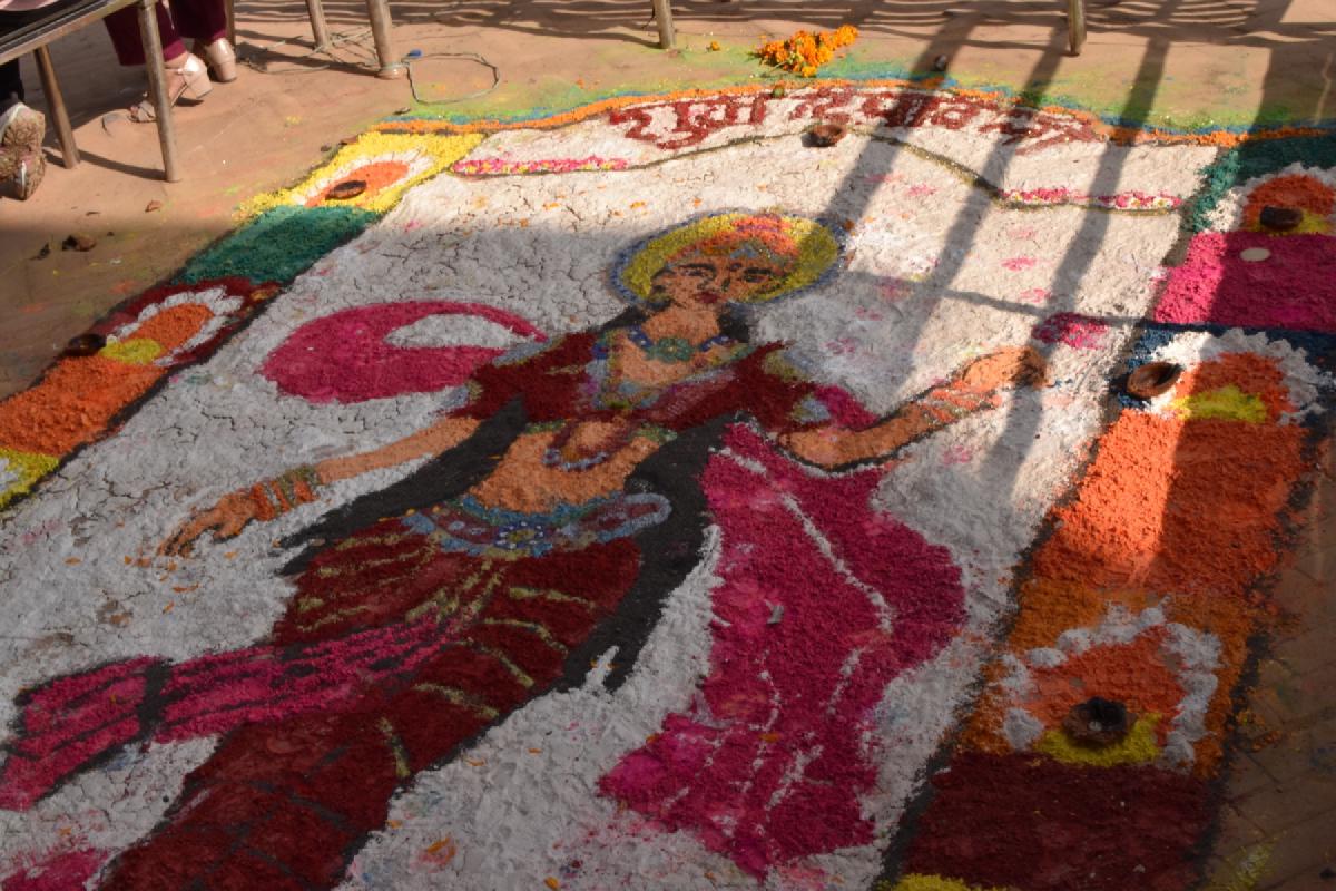 Colored-powder rangoli of a goddess in pink robes laid out on the ground in Nepal, oil lamps set along the border