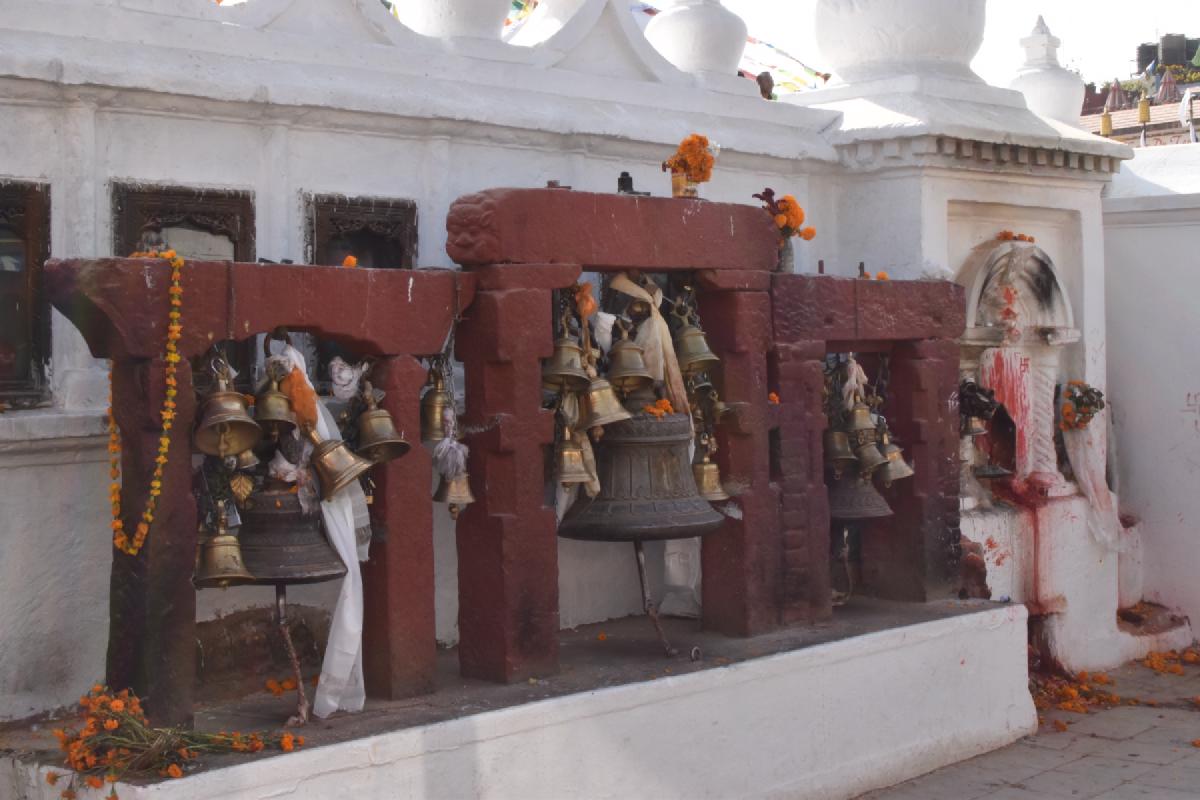 Bronze temple bells hung in red stone frames on a white shrine wall in Nepal, marigold garlands draped around them