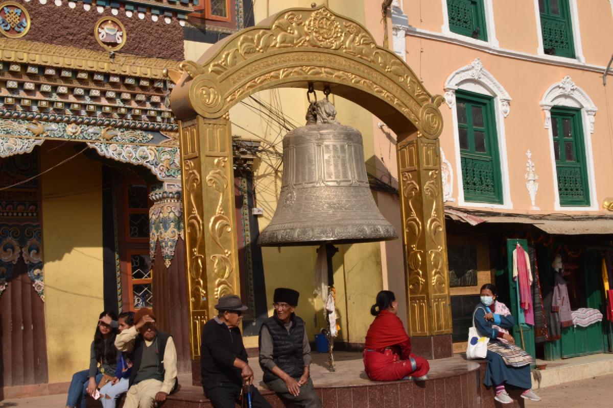 People sitting under a huge engraved silver bell hanging from an ornate golden arch near Boudhanath in Kathmandu