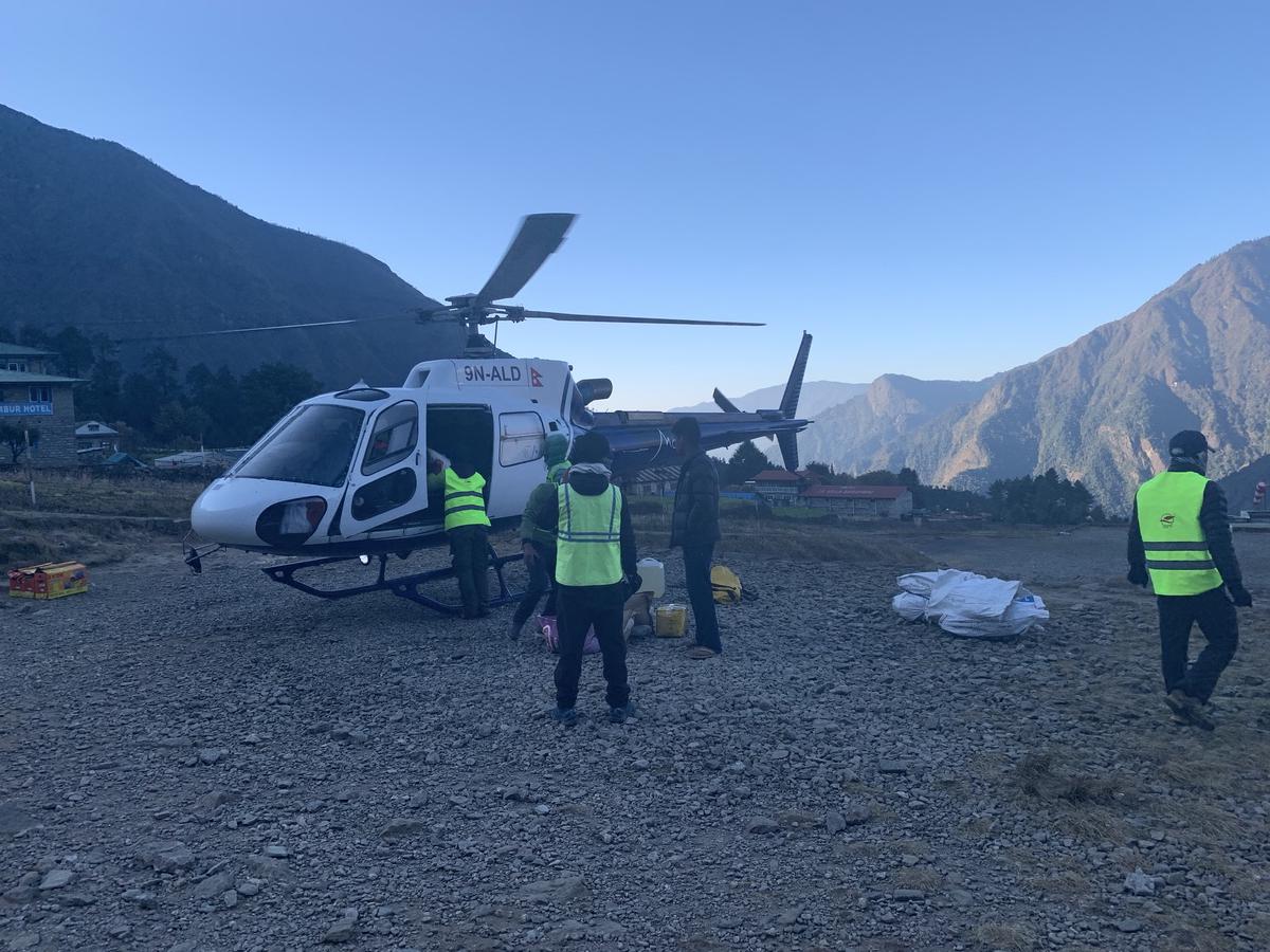 Workers in safety vests loading a helicopter on a gravel pad at Lukla, Nepal, steep forested mountains all around
