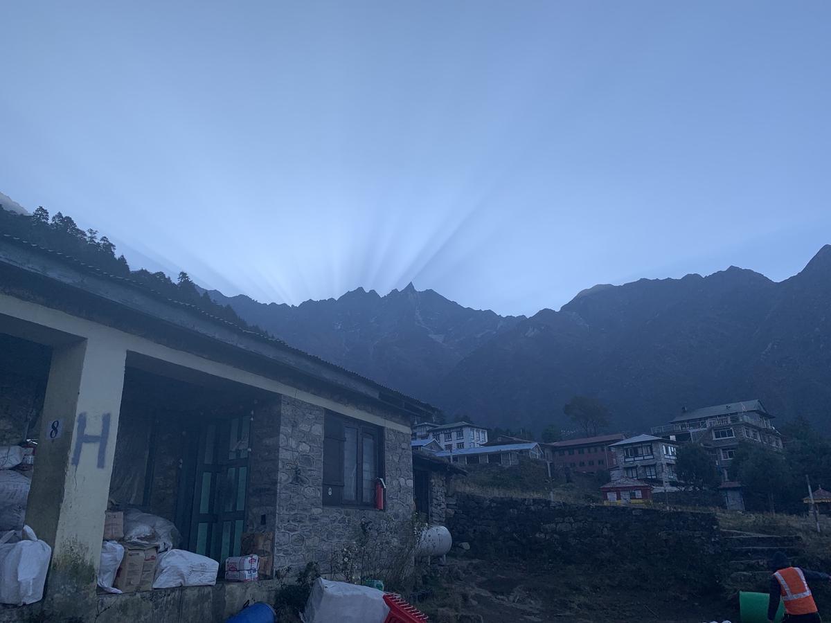 Dawn rays fanning over a dark Himalayan ridge above stone lodges and stacked supplies in Lukla, Nepal