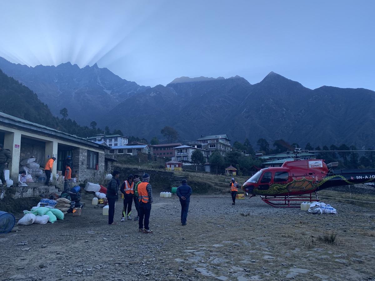 Crews in orange vests staging cargo beside a red helicopter at dawn in Lukla, Nepal, mountains looming in half-light