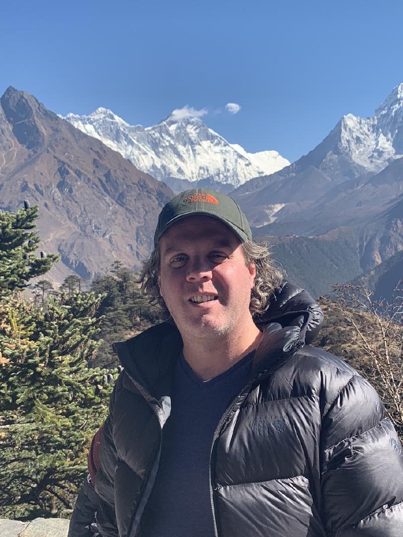 Smiling man in a ball cap and puffy jacket with Everest, Lhotse, and Ama Dablam rising behind him in Nepal