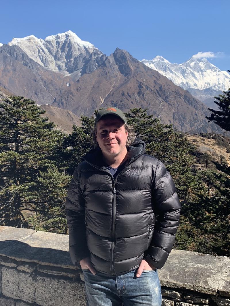 Man in a black down jacket standing at a stone wall in Nepal, pine trees and snowy Himalayan peaks behind him