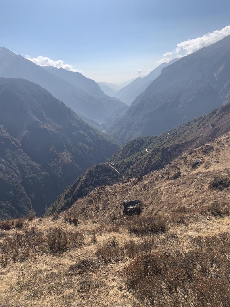 Steep Himalayan valley in Nepal with forested ridges receding into haze, dry golden brush on the slope in the foreground