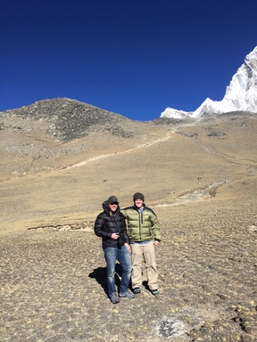 Two men in winter jackets and hats stand on a barren Himalayan slope under deep blue sky, a snowy peak rising behind them